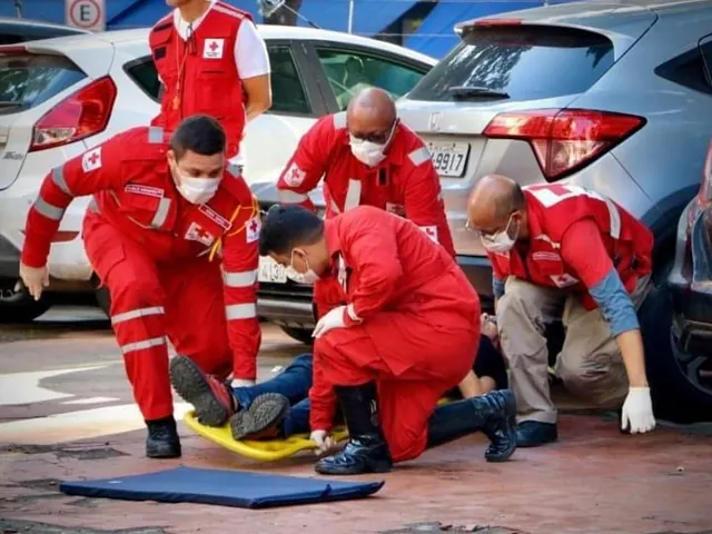 Voluntários de uniforme vermelho fazem atendimento de primeiros socorros a uma pessoa deitada no chão, ao lado de carros